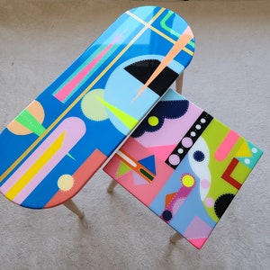 May include: Two colourful abstract painted wooden tables. The larger table is oval shaped and the smaller table is square. Both tables have a blue background with geometric shapes in pink, yellow, green, and black.