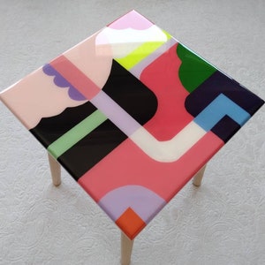 Op de afbeelding: Een vierkante bijzettafel met houten poten en een kleurrijk abstract ontwerp op de bovenkant. Het ontwerp bevat geometrische vormen in roze, rood, groen, blauw, zwart, wit en geel.