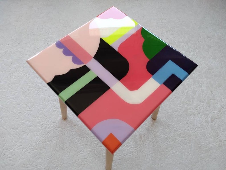 Op de afbeelding: Een vierkante bijzettafel met houten poten en een kleurrijk abstract ontwerp op de bovenkant. Het ontwerp bevat geometrische vormen in roze, rood, groen, blauw, zwart, wit en geel.