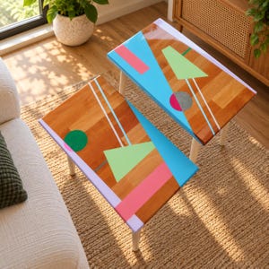 Könnte beinhalten: Zwei rechteckige Beistelltische aus Holz mit weißen Beinen, die eine glänzende Oberfläche mit geometrischen Formen in leuchtendem Blau, Hellgrün, Pink und Lavendel aufweisen, akzentuiert durch grüne und graue Kreise sowie dünne weiße Linien.