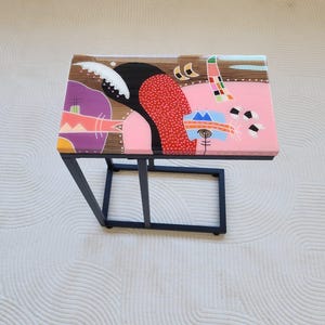 Può includere: Tavolino rettangolare con struttura in metallo nero e un design astratto colorato sul piano. L'opera d'arte presenta forme rosa, rosse, nere e bianche. Il tavolo è posto su uno sfondo bianco strutturato.