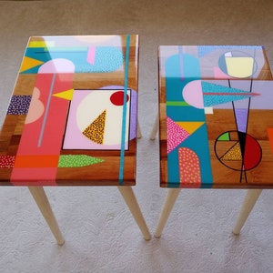 Könnte beinhalten: Zwei Holz-Beistelltische mit geometrischen Mustern, die auf die Tischplatte gemalt sind. Die Tische sind in leuchtenden Farben bemalt, darunter Pink, Blau, Gelb, Grün und Rot. Die Tische haben eine transparente Epoxidharz-Oberfläche.
