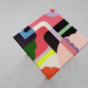Op de afbeelding: Een vierkante tafel met een kleurrijk geometrisch ontwerp. Het tafelblad is gemaakt van hars en heeft een verscheidenheid aan vormen en kleuren, waaronder roze, groen, blauw, geel en zwart.