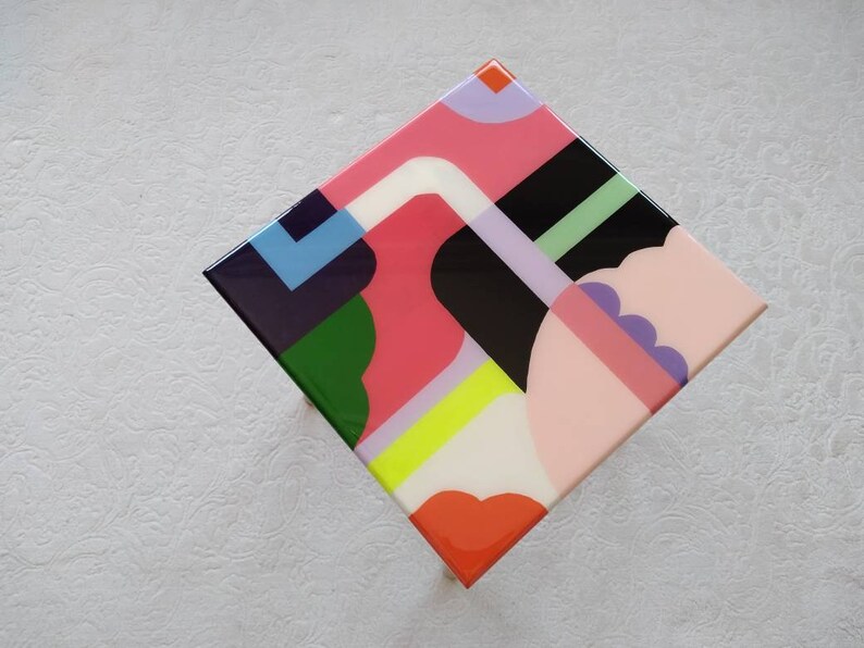 Op de afbeelding: Een vierkante tafel met een kleurrijk geometrisch ontwerp. Het tafelblad is gemaakt van hars en heeft een verscheidenheid aan vormen en kleuren, waaronder roze, groen, blauw, geel en zwart.