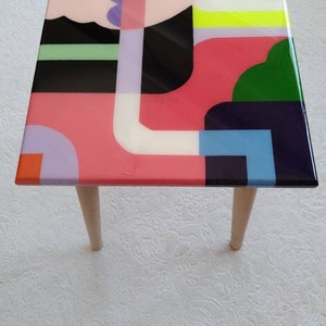 Op de afbeelding: Een vierkante tafelblad met houten poten, met een kleurrijk abstract ontwerp. Het ontwerp bevat geometrische vormen in roze, groen, blauw, zwart, wit en geel.
