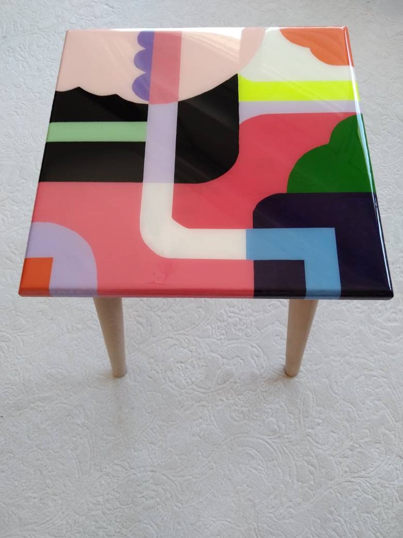 Op de afbeelding: Een vierkante tafelblad met houten poten, met een kleurrijk abstract ontwerp. Het ontwerp bevat geometrische vormen in roze, groen, blauw, zwart, wit en geel.