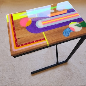 May include: A colorful side table with a geometric design featuring a purple, pink, yellow, and green pattern. The table has a wooden top and a black metal frame.