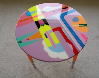May include: A round, pink table with a colorful geometric design. The table has three orange legs with white stripes.