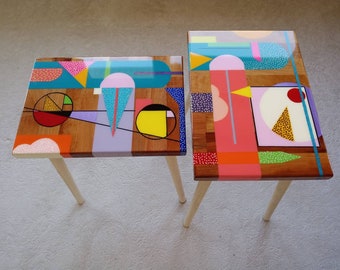 May include: Two wooden side tables with geometric patterns painted in bright colors, including pink, yellow, blue, green, and orange. The tables have wooden legs.