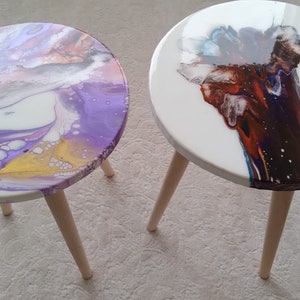 May include: Two round wooden side tables with white tops and wooden legs. The tops are painted with abstract designs in purple, white, gold, and red.