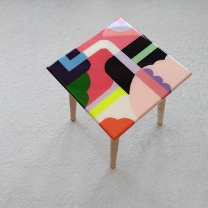 Op de afbeelding: Een vierkante bijzettafel met vier houten poten. Het tafelblad is gemaakt van hars en heeft een kleurrijk abstract geometrisch ontwerp met roze, groene, gele, witte, zwarte en paarse vormen.