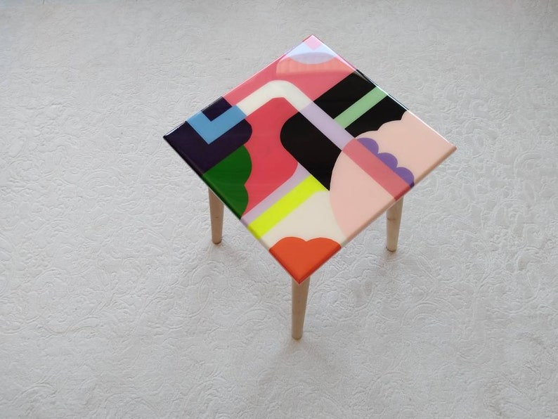 Op de afbeelding: Een vierkante bijzettafel met vier houten poten. Het tafelblad is gemaakt van hars en heeft een kleurrijk abstract geometrisch ontwerp met roze, groene, gele, witte, zwarte en paarse vormen.