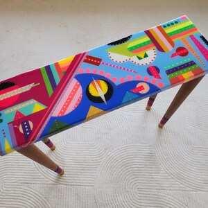 Peut inclure: Une table basse rectangulaire avec un design abstrait coloré. Le plateau présente un mélange vibrant de formes géométriques et de lignes dans des tons de bleu, rose, jaune et vert. La table a quatre pieds en bois.
