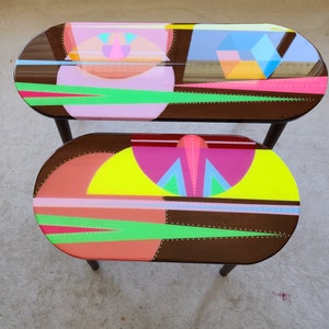 May include: Two oval-shaped coffee tables with black legs. The tabletops are made of dark wood with a clear epoxy finish. The top table has a pink, yellow, and blue geometric design. The bottom table has a red, yellow, and green geometric design.