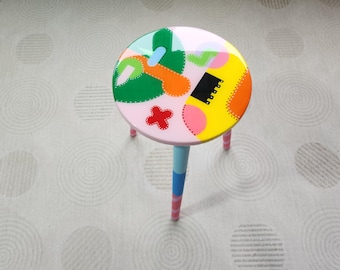 May include: A colorful, hand-painted stool with a pink base and multi-colored geometric designs on the top. The stool has three legs, each painted a different color.