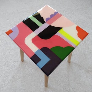 Op de afbeelding: Een kleine vierkante tafel met houten poten en een kleurrijk abstract ontwerp op de bovenkant. Het ontwerp bevat geometrische vormen in roze, groen, blauw, zwart, wit en geel.