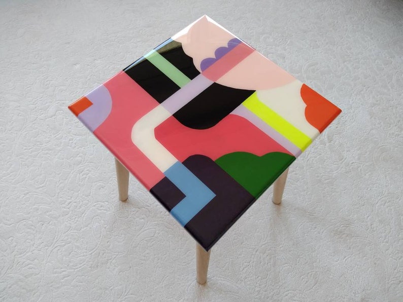 Op de afbeelding: Een kleine vierkante tafel met houten poten en een kleurrijk abstract ontwerp op de bovenkant. Het ontwerp bevat geometrische vormen in roze, groen, blauw, zwart, wit en geel.
