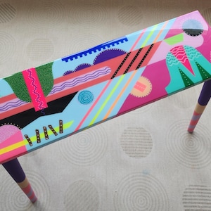 May include: A colourful geometric design painted on a light blue table with three legs. The table is painted in shades of pink, blue, green, yellow, and black.