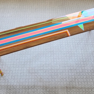 May include: A long wooden bench with a glossy resin finish featuring vibrant neon geometric lines in pink, blue, green, and orange. The surface displays a natural wood grain, and the cylindrical legs are painted in sections of lime green and pink.