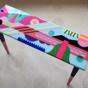 Könnte beinhalten: Ein farbenfroher Tisch mit geometrischem Muster, einer hellblauen Basis und violetten Beinen. Die Tischplatte zeigt eine Vielzahl von Formen und Farben, darunter Pink, Grün, Schwarz und Gelb.