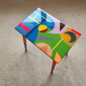 Peut inclure: Petite table rectangulaire au design géométrique vibrant. Le plateau présente un motif coloré de formes bleues, vertes, roses et oranges. Les pieds sont oranges avec des accents rayés. Un meuble unique.