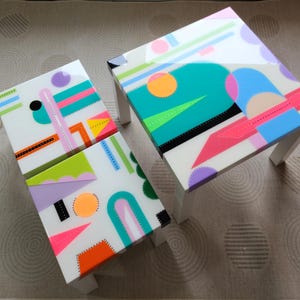 May include: Three white square tables with geometric designs painted on the tops. The designs feature bright colours like pink, orange, green, blue, and yellow. The tables are on a beige carpet with a circular pattern.