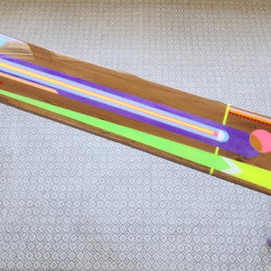 Könnte beinhalten: Ein langer, rechteckiger Couchtisch aus Holz mit einem farbenfrohen, geometrischen Design. Der Tisch hat eine natürliche Holzmaserung mit neongrünen, lila, orangefarbenen und gelben Akzenten. Die Beine sind orange und grau lackiert. Der Tisch ist ca. 152 cm lang.