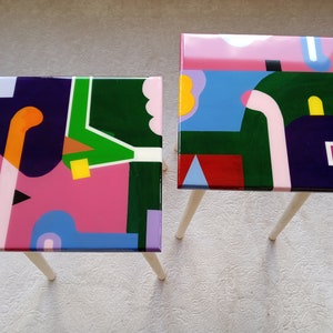 May include: Two colorful abstract painted wooden side tables with white legs. The tabletops feature geometric shapes in pink, green, blue, yellow, red, black, and purple.
