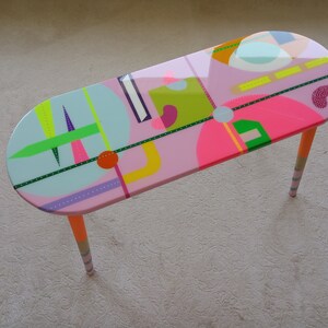 May include: A pink oval coffee table with colorful geometric designs and orange, white, and gray striped legs.