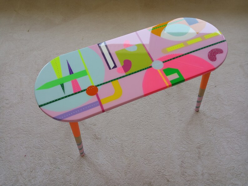 May include: A pink oval coffee table with colorful geometric designs and orange, white, and gray striped legs.
