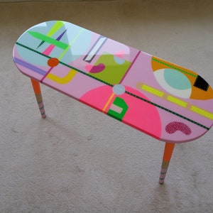 May include: A pink oval coffee table with colorful geometric designs and orange and white striped legs.