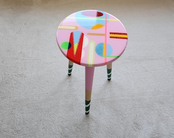 May include: A small, round side table with a pink top featuring colorful abstract shapes. The table has three legs, each with a pink upper section and a lower section painted with green and white stripes. The table's top is approximately 15 inches in diameter.