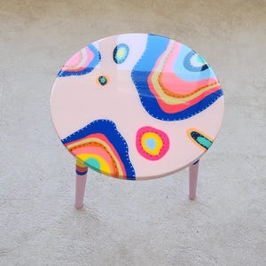 May include: A round, pink side table with three legs. The tabletop features a colorful abstract design with blue, yellow, pink, and orange shapes. The legs are painted pink with colorful stripes near the top.
