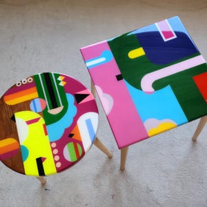 May include: Two colourful abstract geometric design coffee tables with wooden legs. The larger table has a pink, green, blue, and white design. The smaller table has a brown, yellow, red, and green design.