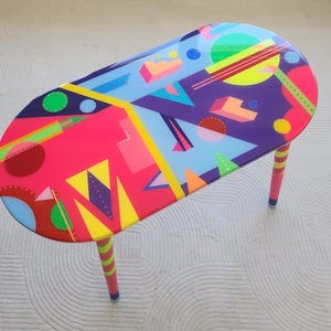 Op de afbeelding: Een ovale salontafel met een levendig geometrisch ontwerp. Het tafelblad heeft een mix van roze, paarse, gele en blauwe vormen. De poten zijn roze met gele strepen. De tafel is ongeveer 122 cm lang.