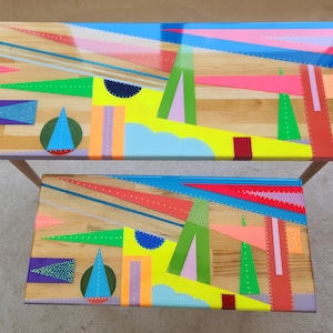May include: Two wooden tables with geometric patterns painted in bright colours, including yellow, green, blue, pink, and red. The tables are stacked on top of each other.