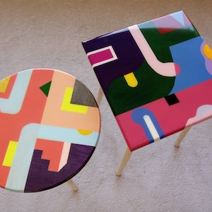 May include: Two colourful geometric patterned end tables with wooden legs. The tabletops are painted with abstract shapes in shades of pink, blue, green, yellow, and purple.