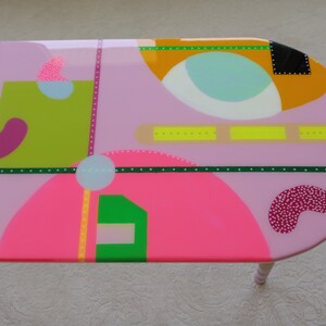 May include: A pink oval table with a geometric design featuring circles, squares, and rectangles in various colors, including green, yellow, pink, and blue.