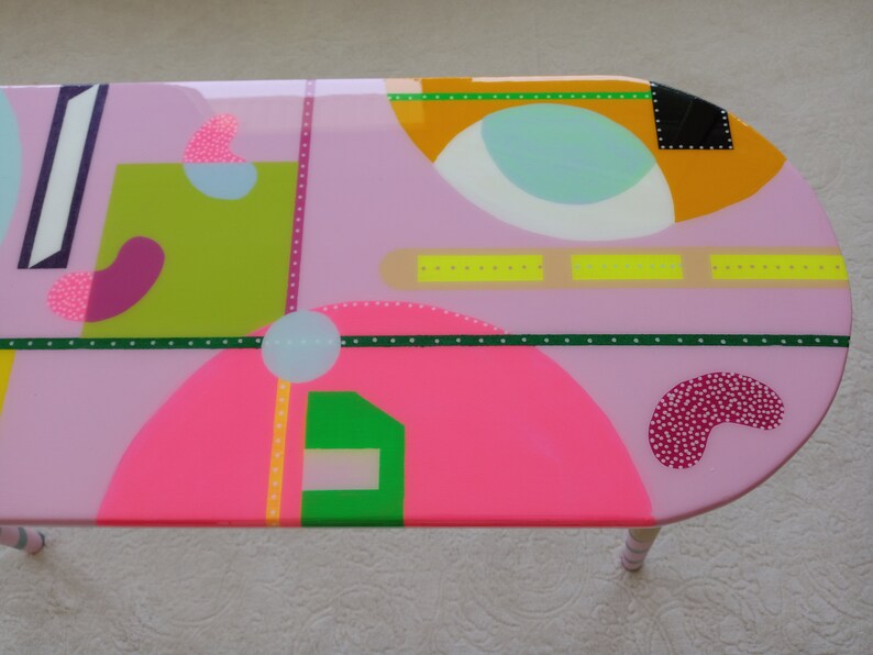 May include: A pink oval table with a geometric design featuring circles, squares, and rectangles in various colors, including green, yellow, pink, and blue.