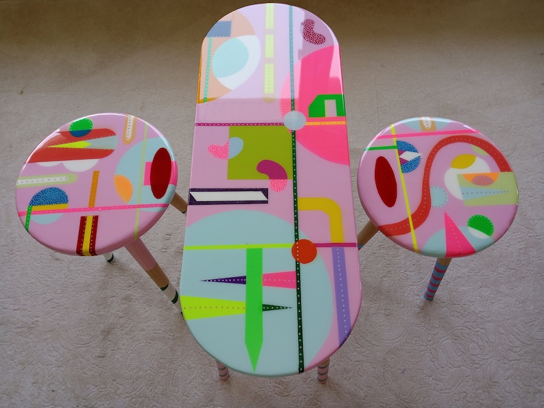 May include: A pink oval table with a geometric design in bright colors, including green, yellow, blue, and red. The table is accompanied by two matching pink round stools with similar geometric designs.