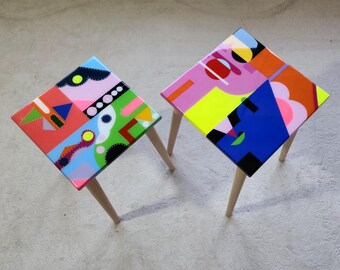 May include: Two colorful geometric patterned wooden stools with light wood legs. The stools are painted with bright colors, including pink, blue, green, yellow, and orange.