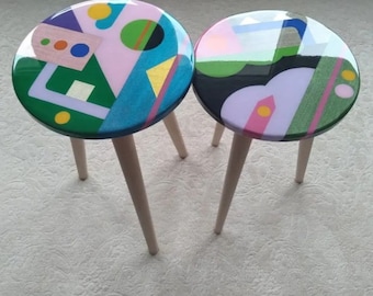 May include: Two small, round stools with colorful abstract designs on the tops. The designs feature geometric shapes in pink, green, blue, and gold. The stools have light-colored wooden legs.