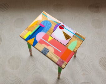 May include: A small wooden table with a colorful geometric design painted on the top. The table has three legs painted in a light green color.