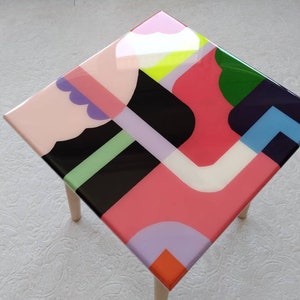 Op de afbeelding: Een vierkante houten tafel met een kleurrijk geometrisch ontwerp op de bovenkant. Het ontwerp bevat roze, groene, blauwe, zwarte, witte en paarse vormen.