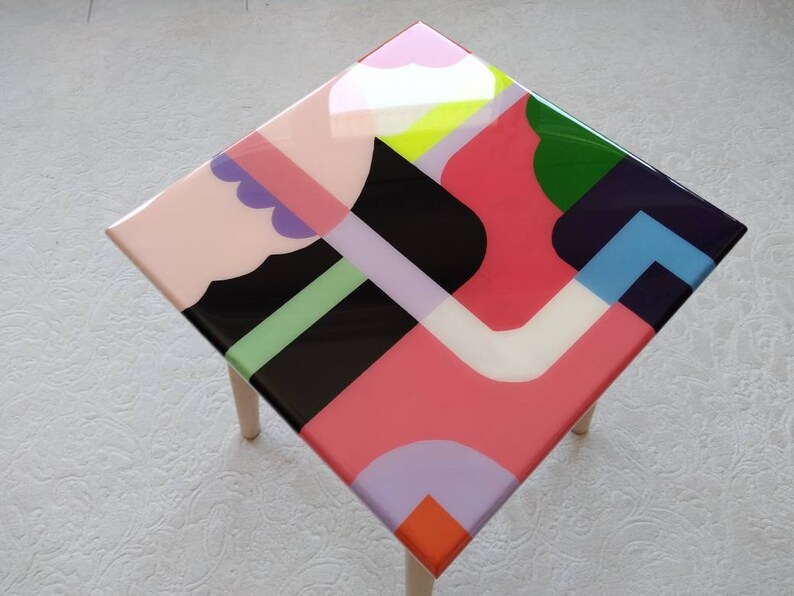 Op de afbeelding: Een vierkante houten tafel met een kleurrijk geometrisch ontwerp op de bovenkant. Het ontwerp bevat roze, groene, blauwe, zwarte, witte en paarse vormen.