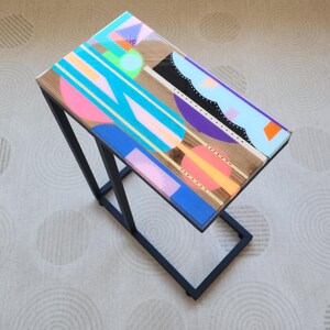 May include: A black metal side table with a wooden top painted with a colorful geometric design. The design features blue, pink, yellow, and green shapes.