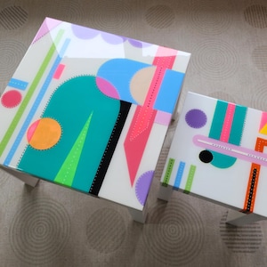 May include: Two white tables with geometric designs in bright colours, including pink, blue, green, orange, and yellow. The larger table has a more complex design with a curved shape, while the smaller table has a simpler design with straight lines.