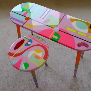 K&ouml;nnte beinhalten: Ein rosa ovaler Tisch mit bunten geometrischen Mustern und ein passender rosa Hocker mit &auml;hnlichen Mustern. Tisch und Hocker haben Holzbeine mit bunten Streifen.