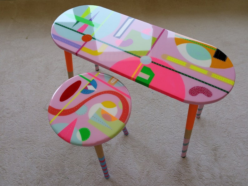 K&ouml;nnte beinhalten: Ein rosa ovaler Tisch mit bunten geometrischen Mustern und ein passender rosa Hocker mit &auml;hnlichen Mustern. Tisch und Hocker haben Holzbeine mit bunten Streifen.