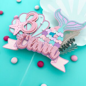May include: A layered mermaid-themed cake topper with the name "Olivia" and the number "3". The topper features a pink glitter finish, a starfish, and a mermaid tail in shades of purple and blue. Decorative elements include small pearls and a banner.